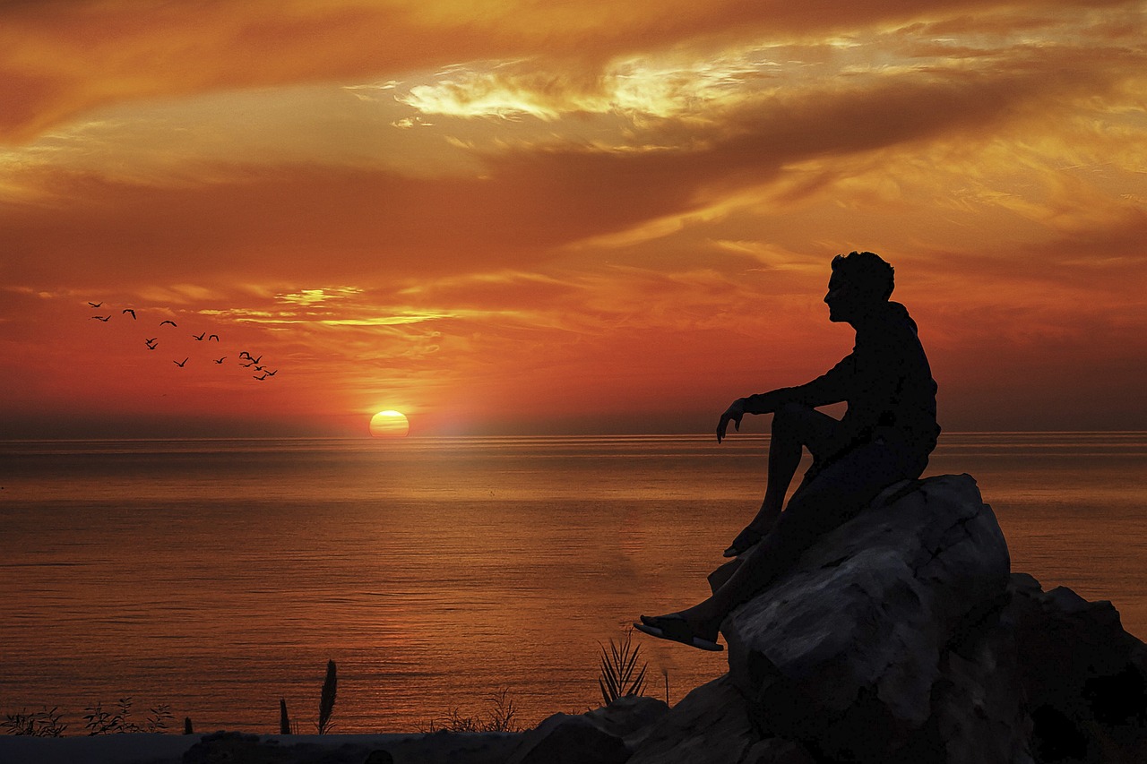 Man on a rock at end of day before the setting sun and a bay